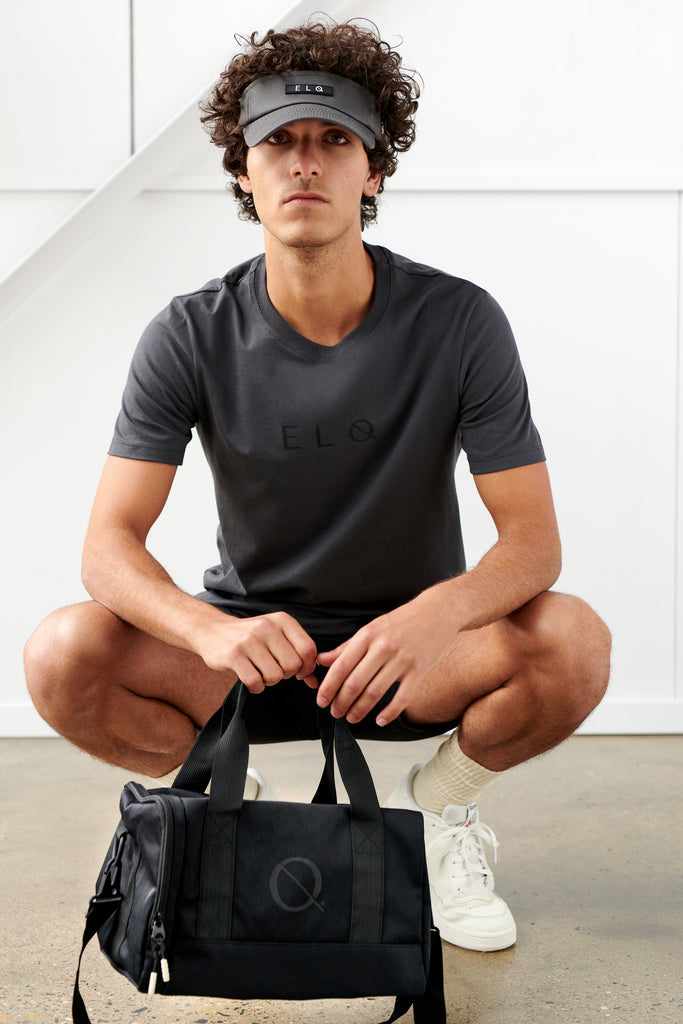 Male model wearing mens black tee, black visor hat and black small crossbody bag duffle